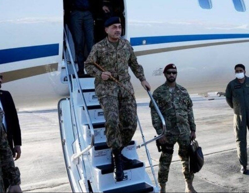 A man in military camouflage uniform descends the airstair of a white aircraft, carrying a wooden baton, while soldiers and another person stand on the tarmac nearby.