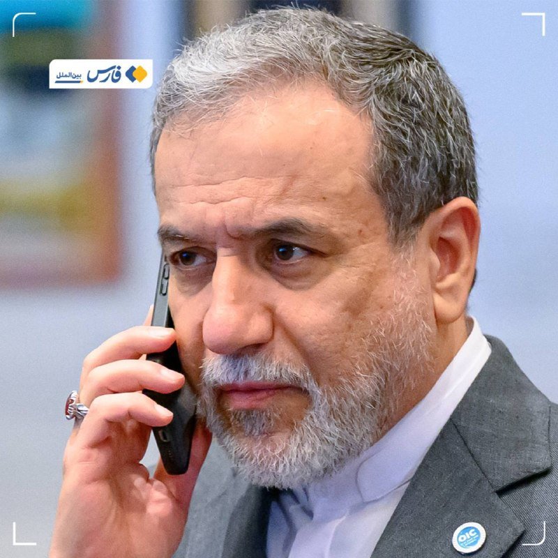 A gray-bearded man in a dark suit holds a smartphone to his ear, speaking with a serious expression.