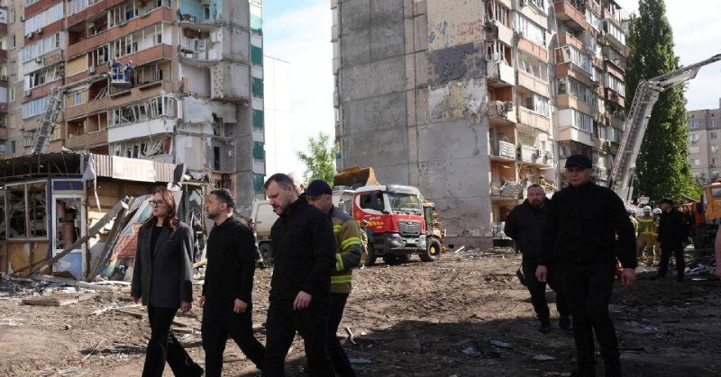 Several people walk through a debris-strewn area in front of a heavily damaged multi-story apartment building, with a fire truck and construction equipment visible in the background.