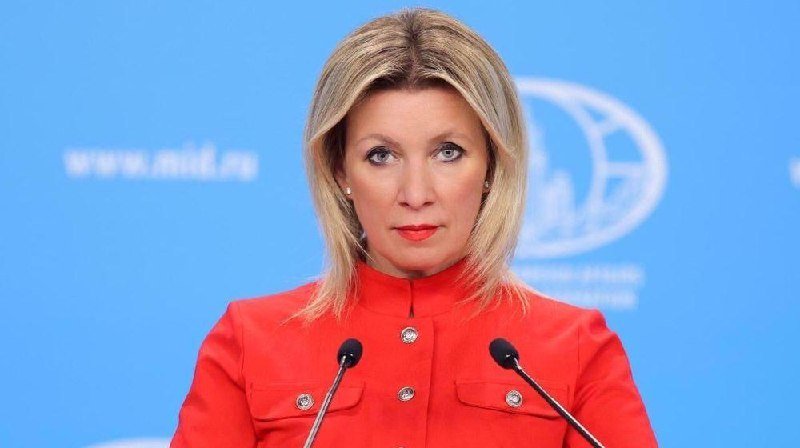 A blonde woman in a red high-collared jacket stands at two microphones against a blue backdrop displaying a partial website URL and a circular emblem.