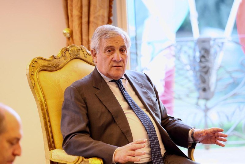 An older man in a dark suit and tie sits in an ornate yellow chair, gesturing with both hands while speaking in a formal room.