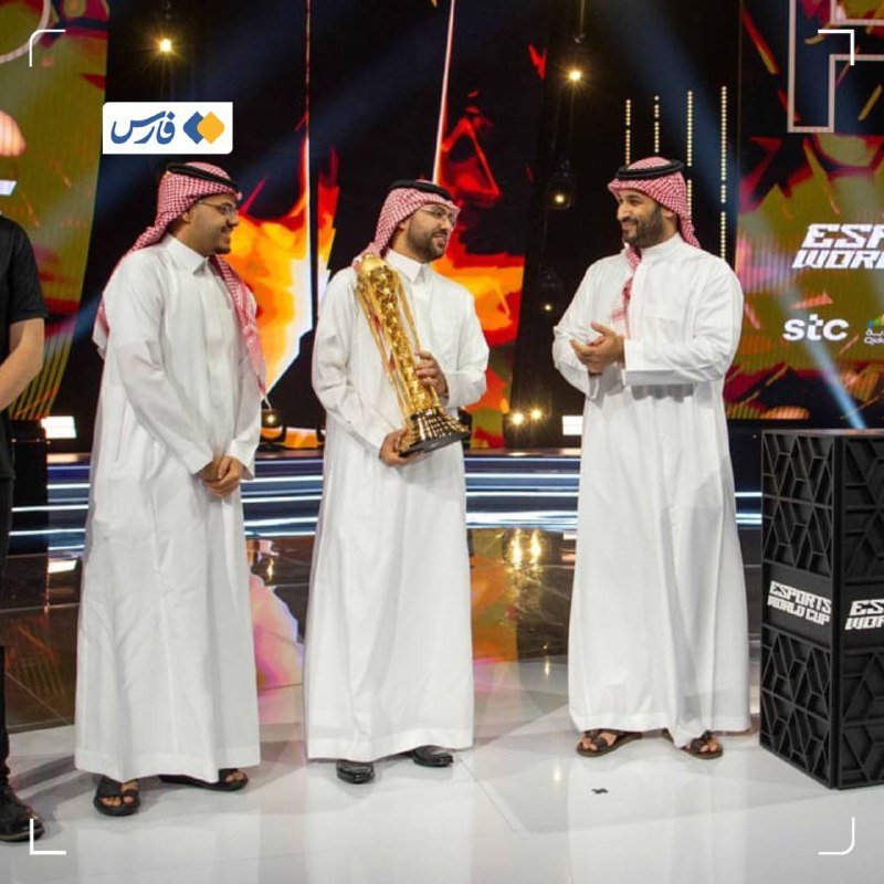 Three men in traditional white thobes and red-checkered keffiyehs stand on an illuminated stage, with one holding a large golden trophy in front of an "Esports World Cup" backdrop.