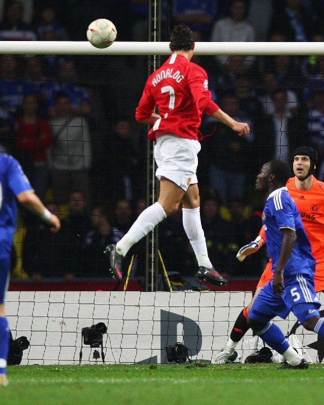 A soccer player wearing a red jersey with "RONALDO 7" jumps in front of the goal as a ball flies overhead, with a goalkeeper and a defender in blue nearby.