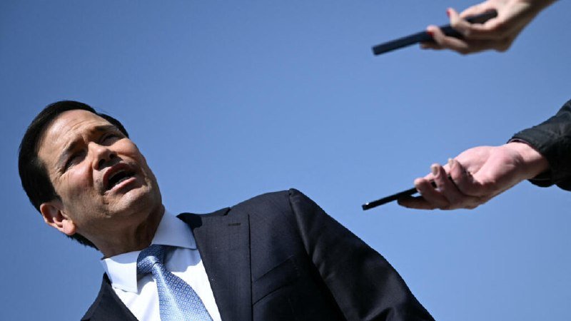 A man in a dark suit and blue tie speaks while two hands holding recording devices extend toward him against a clear blue sky.