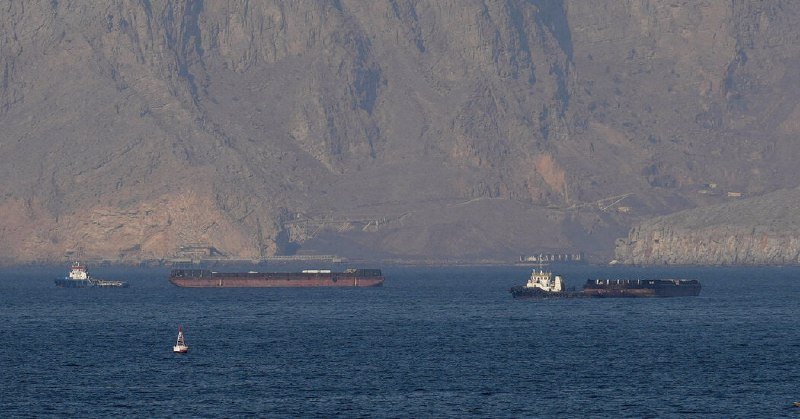 Several large cargo ships and tugboats float on calm blue waters in front of a towering, rugged rocky coastline.