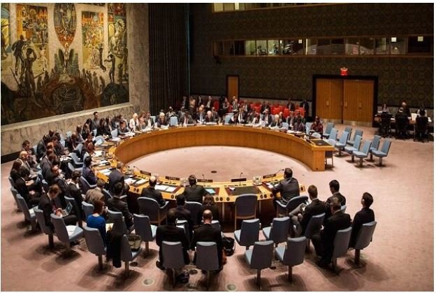Delegates in business attire are seated around a large circular conference table in a formal assembly hall featuring a prominent mural on the wall.