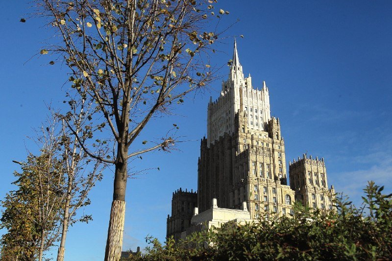A tall, ornate stone skyscraper with a central spire rises against a clear blue sky, partially framed by a sparse, autumn-leaved tree and evergreen shrubs in the foreground.