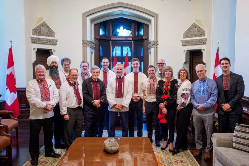 A group of men and women in traditional embroidered Ukrainian shirts and formal attire pose together in an ornate room with Canadian flags and a stone-carved doorway in the background.