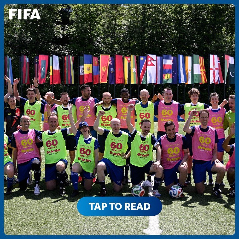 Two rows of people wearing neon yellow and pink "60 Be Active" bibs kneel and stand smiling on grass, with a row of national flags and soccer balls displayed behind them.