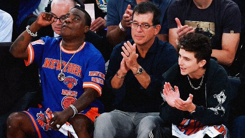 A man in a New York Knicks jersey sits courtside alongside two clapping men at a basketball game.
