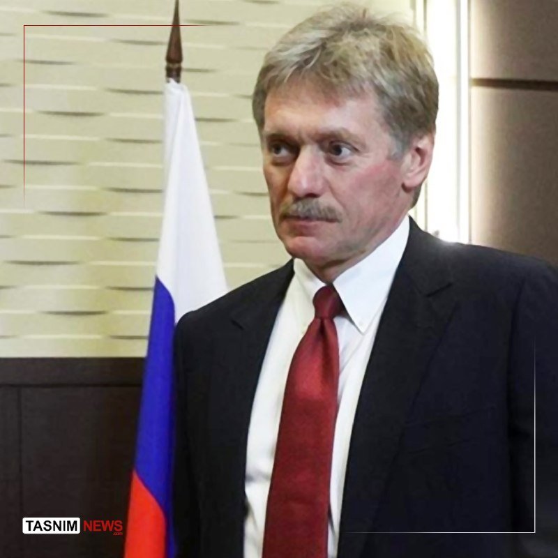 A gray-haired, mustached man in a dark suit and red tie stands beside a white, blue, and red flag.