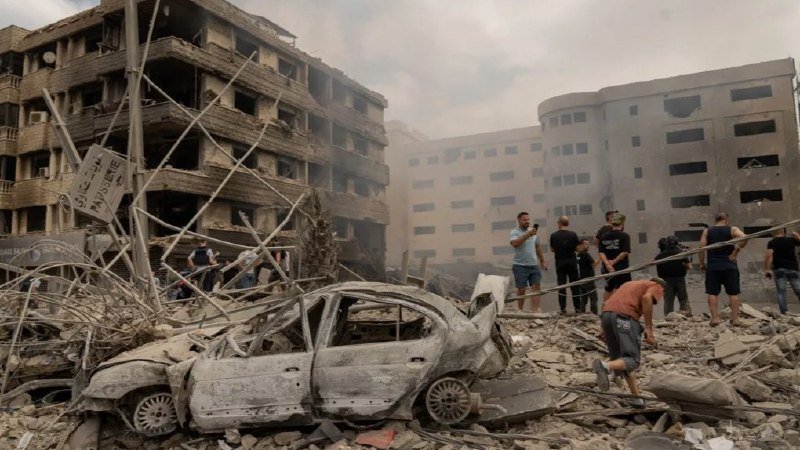 Burnt-out cars and damaged apartment buildings stand amid rubble as people gather in the dusty aftermath of an apparent blast or strike.