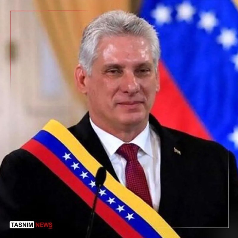 A gray-haired man in a dark suit and red tie stands at a podium wearing a yellow, blue, and red presidential sash with white stars, with flags visible behind him.