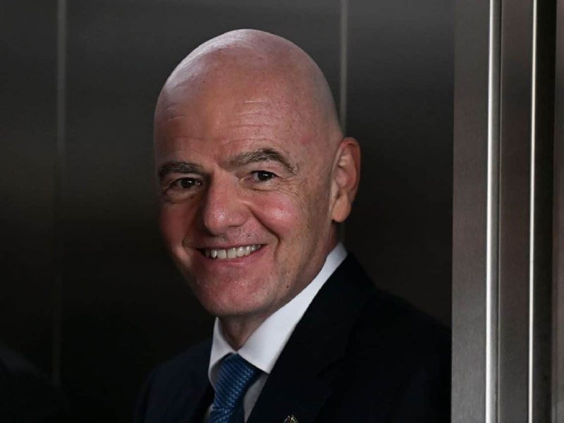 A smiling bald man in a dark suit, white shirt, and blue tie stands against a dark background near a metallic frame.