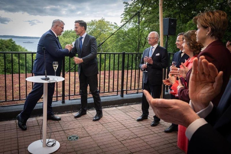 Two men in suits shake hands on an outdoor terrace overlooking water, while others in formal attire applaud nearby.