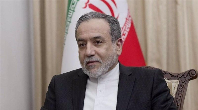 A gray-bearded man in a dark suit and white shirt sits before an Iranian flag.