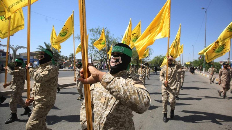 Masked individuals in camouflage uniforms and green headbands march down a street carrying yellow flags.