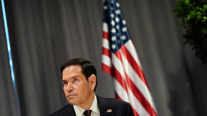 A man in a dark suit with an American flag pin sits before an American flag and gray curtain backdrop.