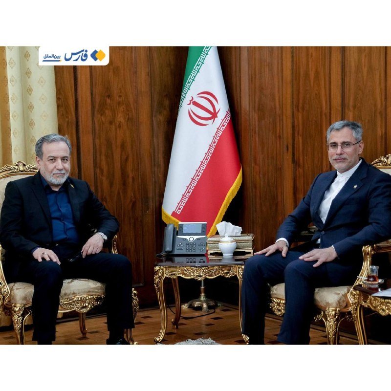 Two men in dark suits sit in ornate gold-trimmed chairs on either side of an Iranian flag, with a telephone and tissue box on a small table between them.