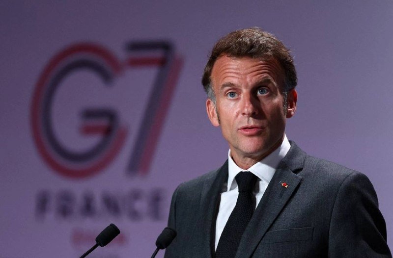 A man in a dark suit and black tie speaks at a podium with microphones, with a blurred "G7 FRANCE" logo visible behind him.