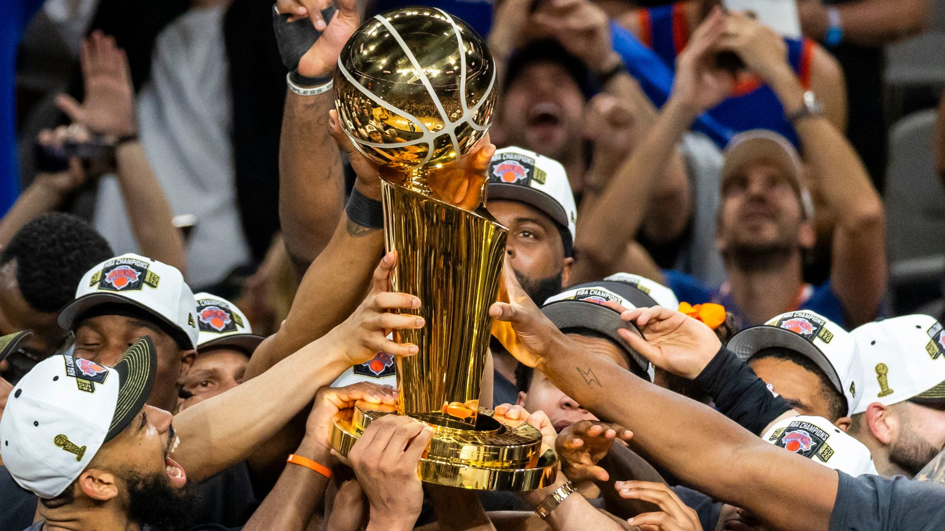 Basketball players wearing "NBA Champions" caps jointly hoist a golden basketball-shaped trophy amid a cheering crowd.