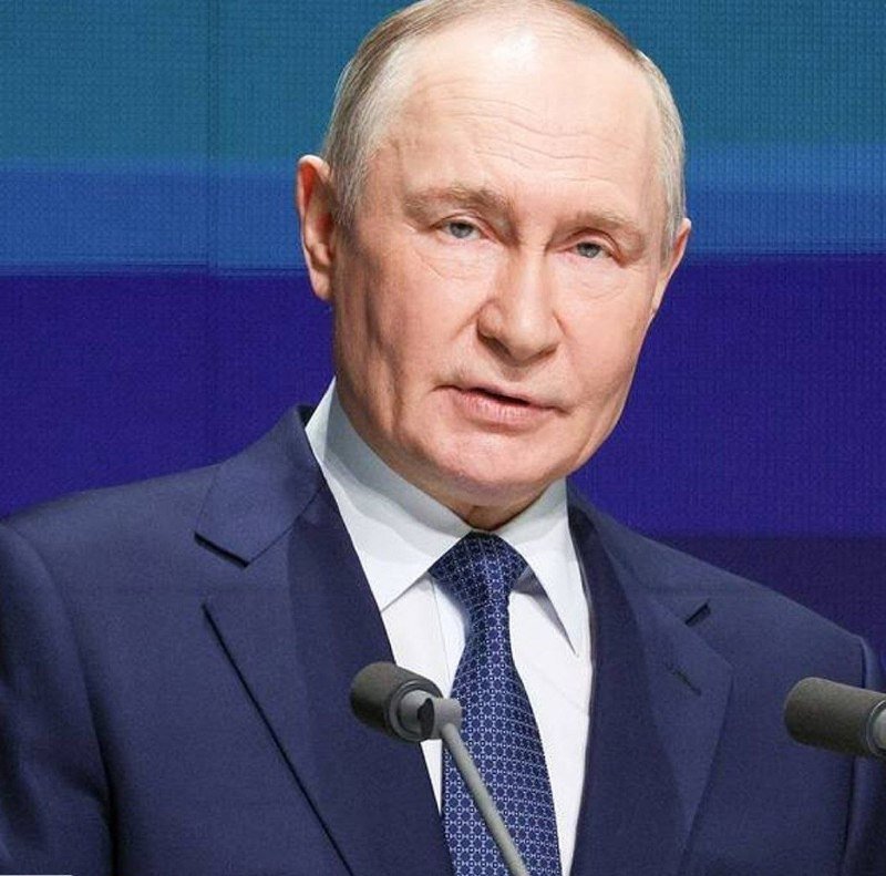 An older man with light hair in a dark suit and patterned blue tie speaks at a podium with microphones against a blue backdrop.
