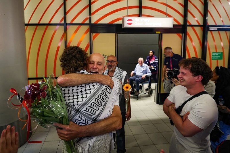 Two people embrace in a hug, one holding a bouquet of flowers, while onlookers smile in a terminal with an "Entry" sign overhead.
