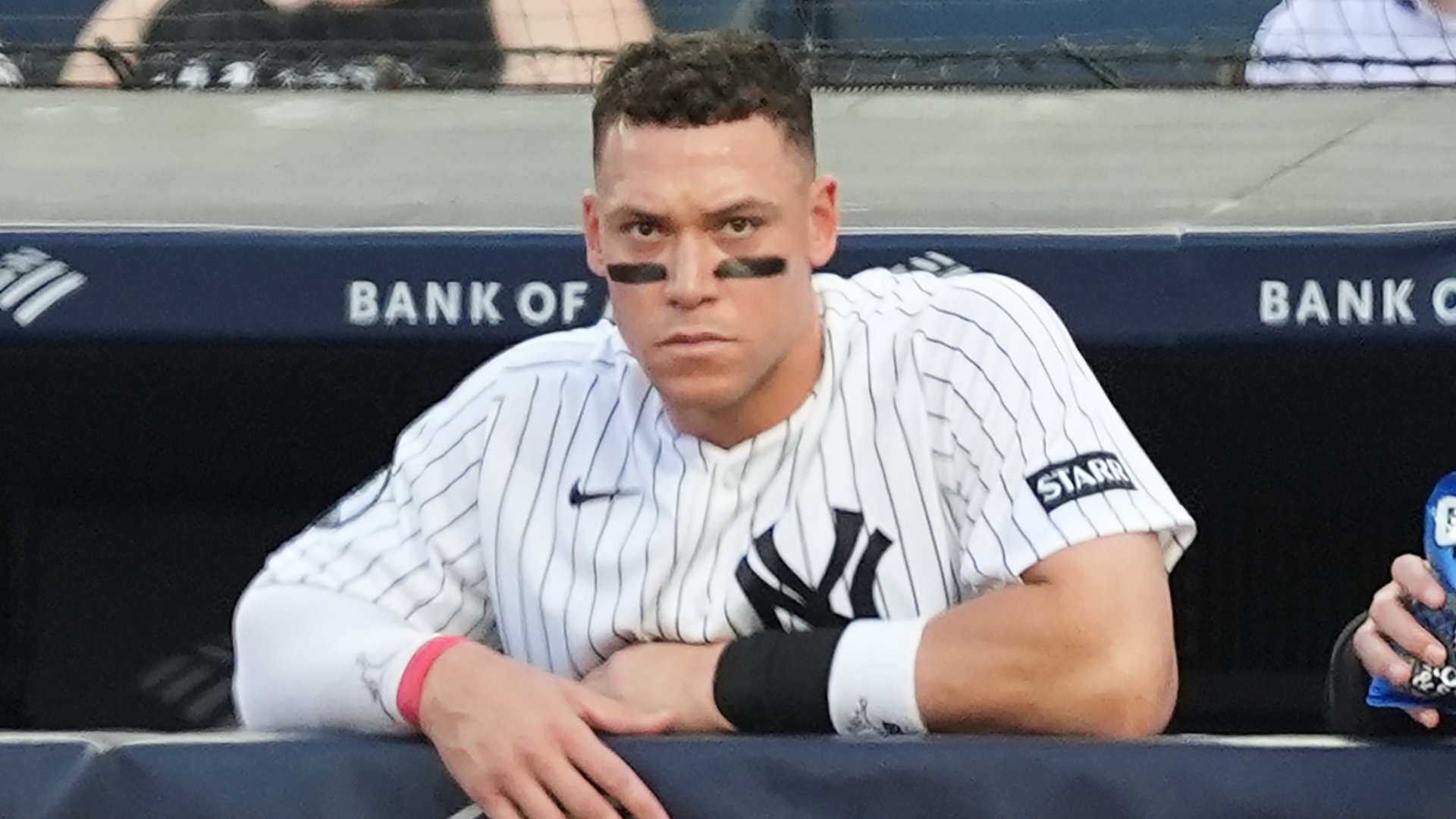 A baseball player in a white pinstriped Yankees uniform with eye black sits in the dugout, leaning on the railing during a game.