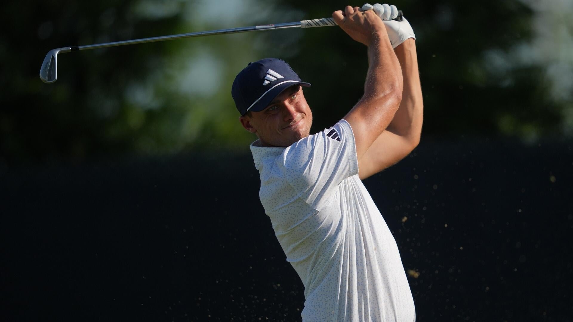 A golfer in a white patterned shirt and dark cap completes a swing, holding an iron raised overhead against a dark, blurred background.