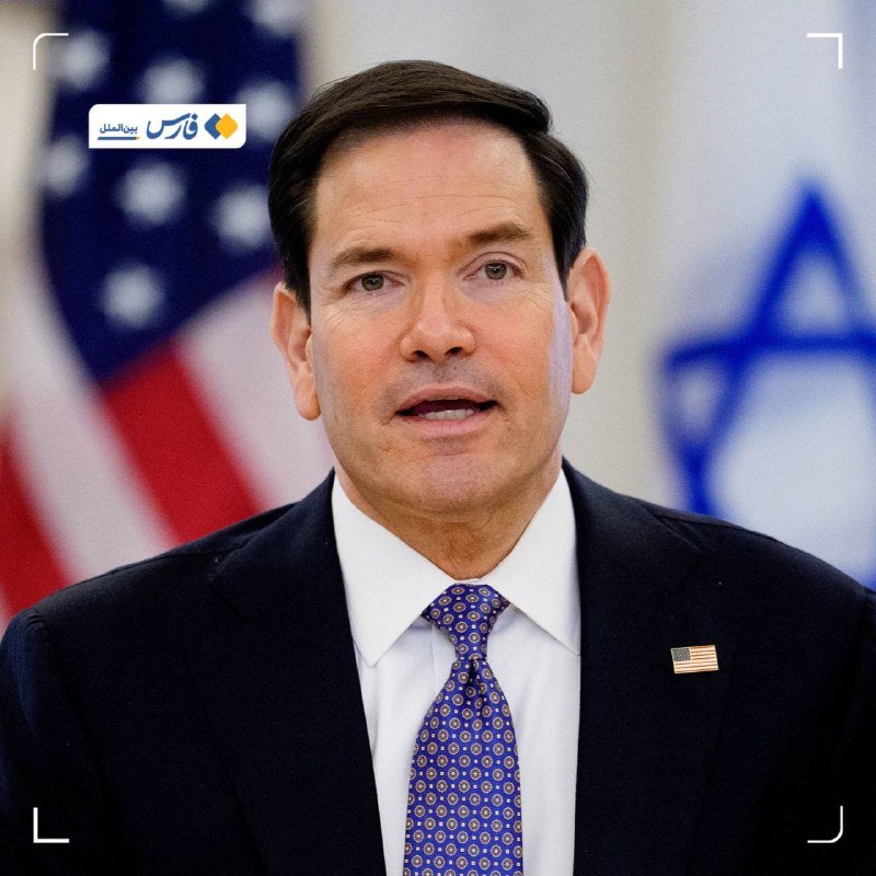 A man in a dark suit, white shirt, and blue patterned tie speaks, with an American flag and a blue-and-white emblem visible in the background.