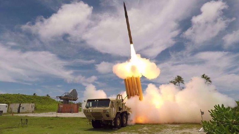 A missile launches from a mobile military launcher on a grassy coastal site, with a radar unit and shipping container visible nearby under a partly cloudy sky.
