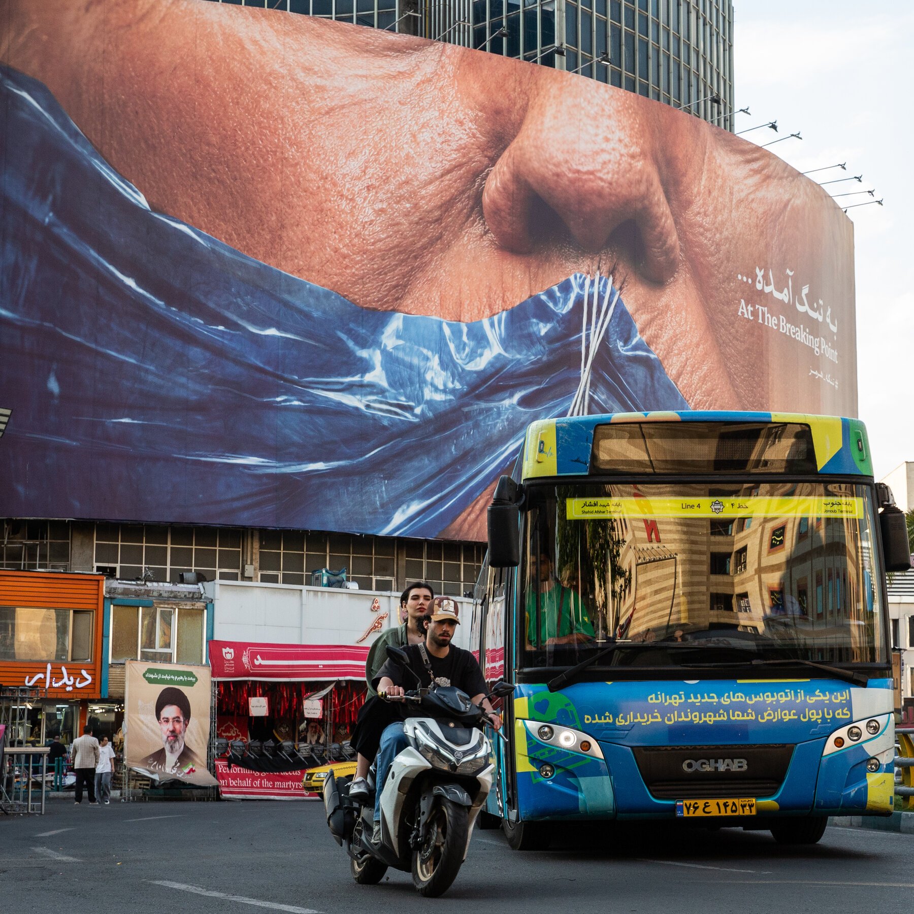 A blue city bus and two people on a motorcycle travel down a street, passing beneath a massive billboard displaying a large face and the text "At The Breaking Point."