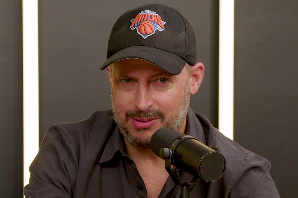 A bearded man wearing a New York Knicks cap and gray shirt speaks into a black microphone against a dark background with vertical light strips.