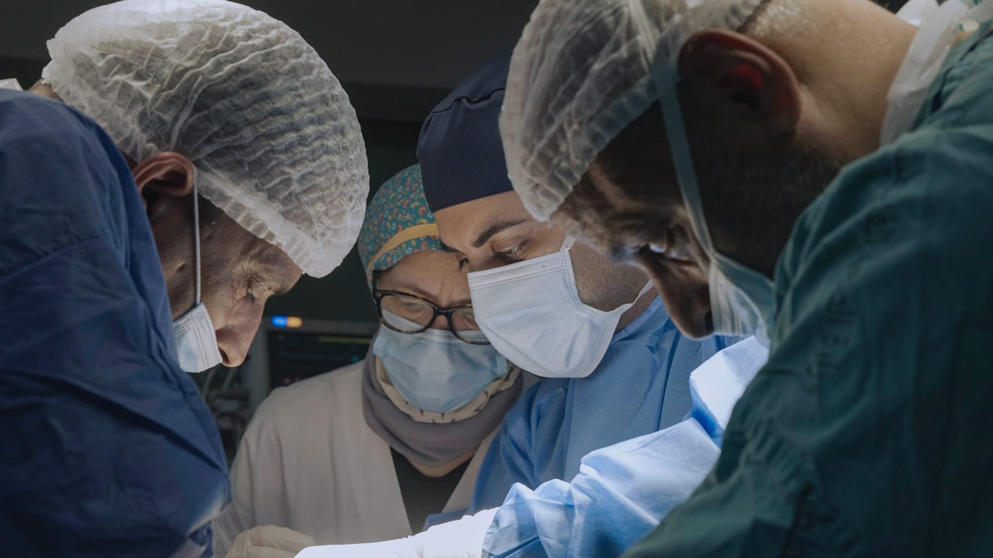 Four surgeons in scrubs, surgical caps, and face masks lean in closely, focused on a procedure in an operating room.