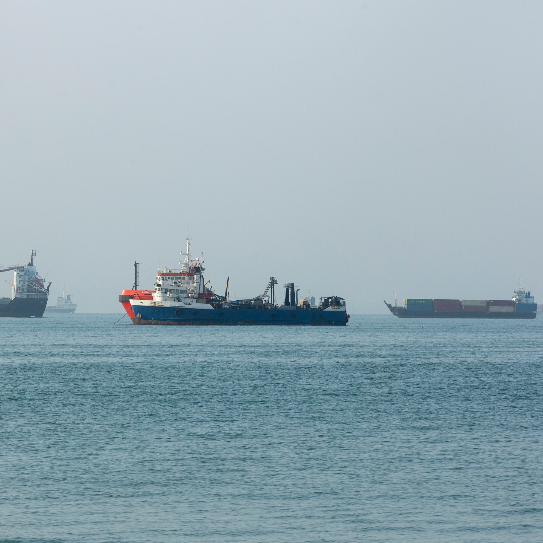 Three large ships, including a central blue-and-red vessel, a cargo ship to the left, and a container ship to the right, sit anchored on calm gray-blue water under a hazy sky.
