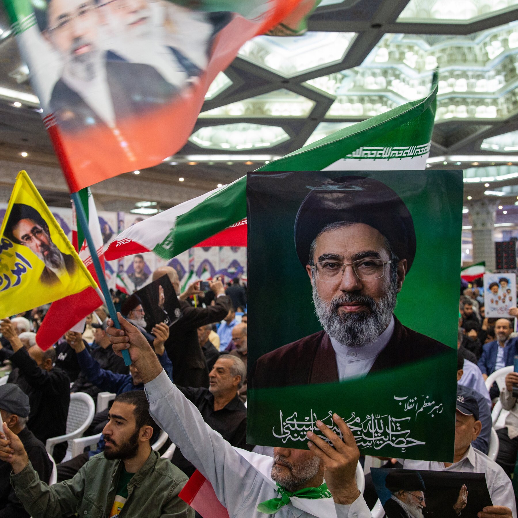 Crowd at indoor rally waves Iranian flags and holds up a portrait of a bearded cleric in black turban alongside yellow and green banners with Persian script.