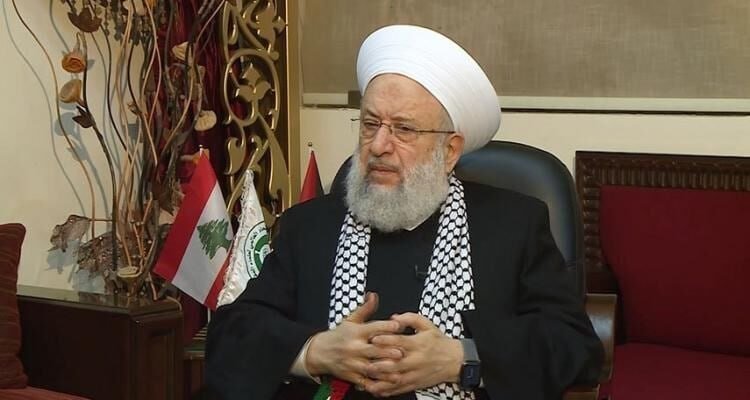 A bearded man wearing a white turban and black-and-white checkered scarf sits with folded hands, beside a Lebanese flag and another emblem flag.
