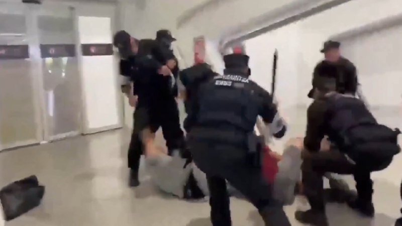 Uniformed officers in tactical gear restrain individuals on the floor of a tiled hallway.