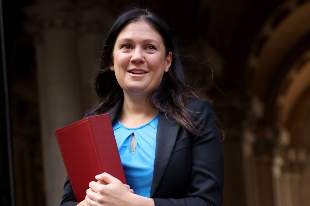 A smiling woman with long dark hair, wearing a black blazer over a blue top, holds a red folder outside a stone-columned building.