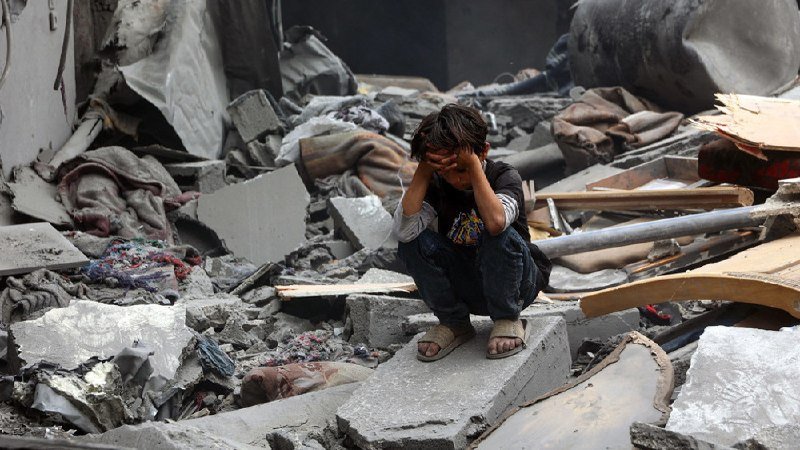 A young child sits on a concrete slab amid extensive rubble, debris, and scattered blankets, covering their face with both hands.
