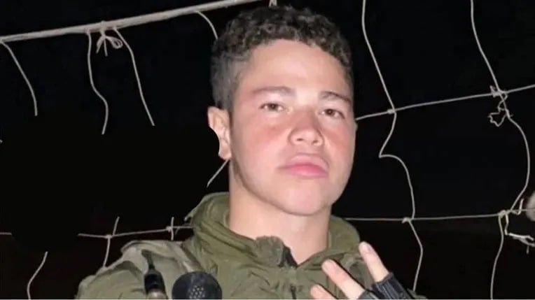 A young man with short dark hair wearing a green hooded jacket is photographed at night, standing in front of a chain-link or wire fence.