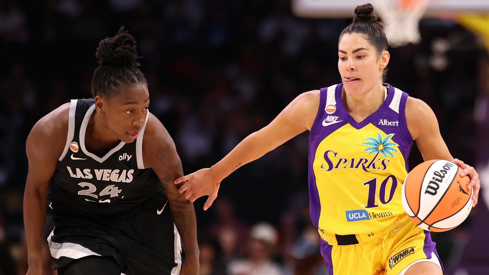 A Sparks player in a yellow and purple #10 jersey dribbles a Wilson basketball while being defended by an Aces player wearing a black #24 Las Vegas jersey.