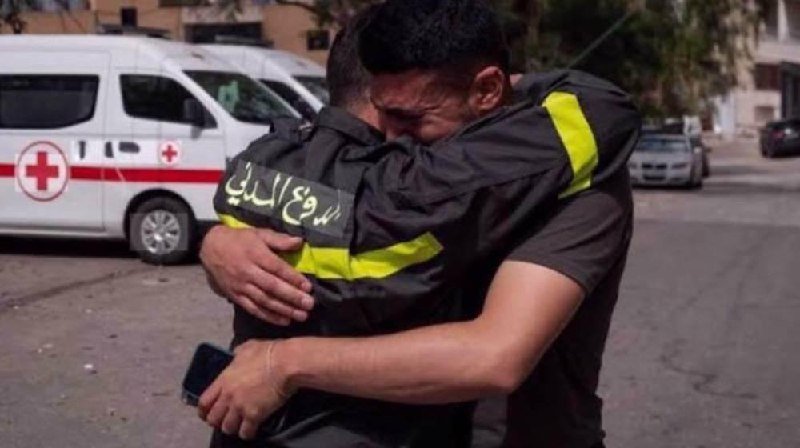 Two men in dark uniforms with reflective yellow stripes embrace tightly on a street, with a white Red Cross ambulance and a parked car visible in the background.