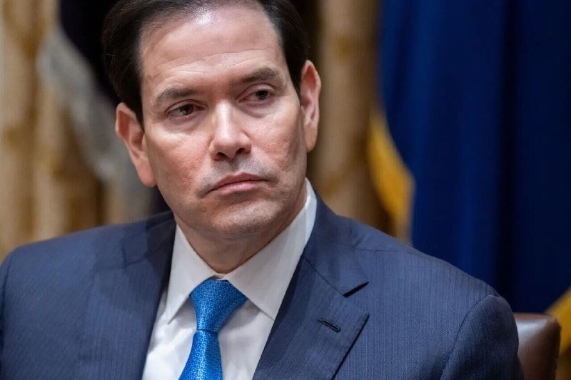 A man in a dark blue suit and blue tie looks to the side with a serious expression, seated in front of blue and gold draped curtains.