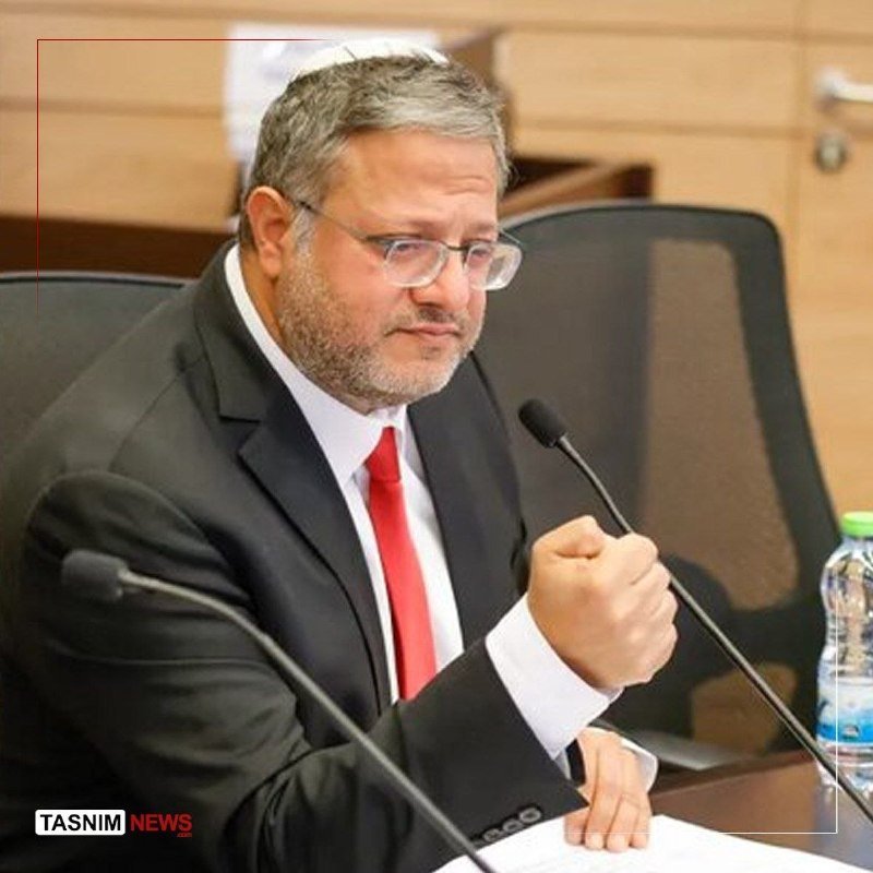 A gray-bearded man in a dark suit, white shirt, red tie, and glasses gestures with his fist while seated at a table with microphones.