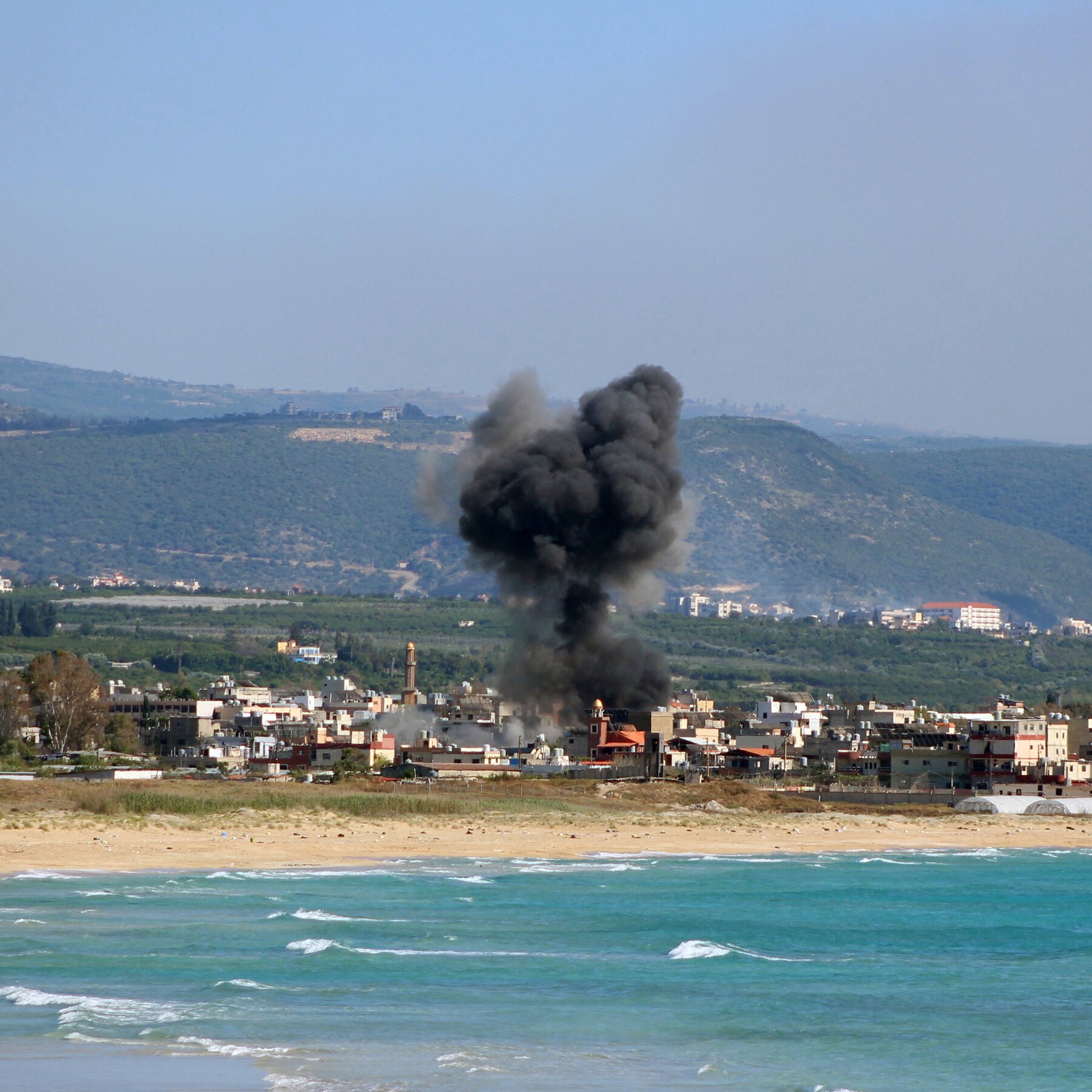 A large plume of black smoke rises above a coastal town of low-rise buildings, with a sandy beach and ocean waves in the foreground and green hills behind.