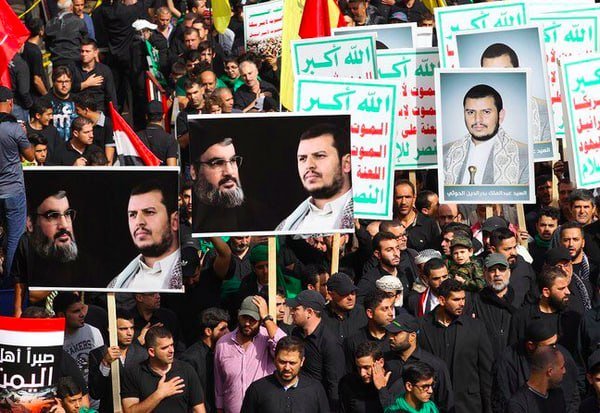 A large crowd of people, many dressed in black, hold up posters featuring portraits of men and Arabic-script signs during a daytime gathering outdoors.