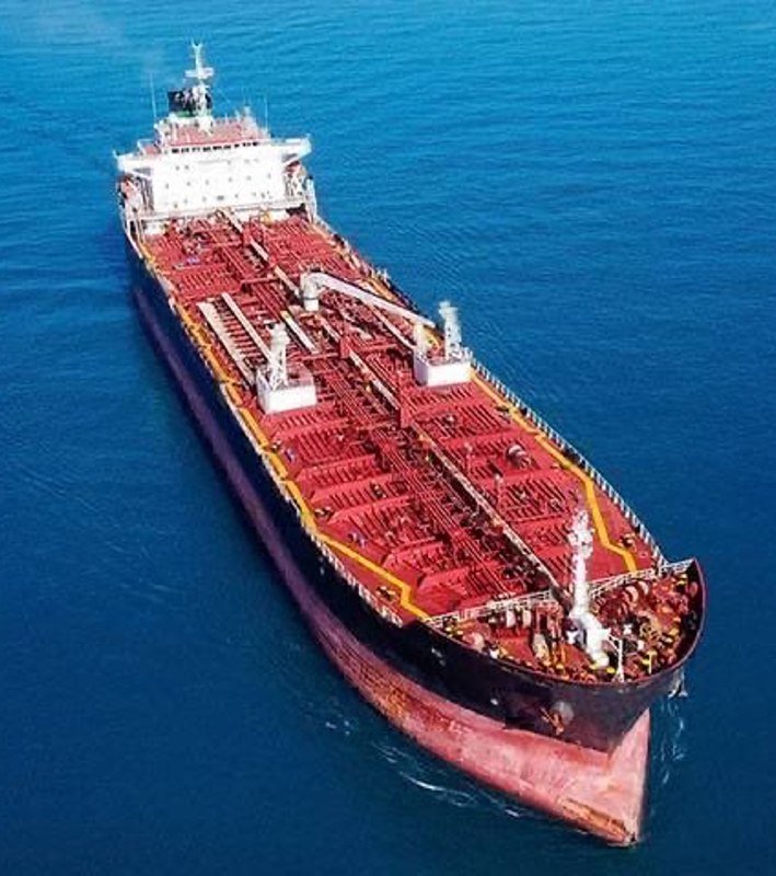 An aerial view shows a large red-and-black oil tanker with a white superstructure sailing on deep blue open water.