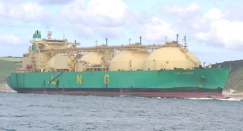 A large green and beige LNG tanker ship sails on the water beneath a cloudy sky, with green hills visible in the background.