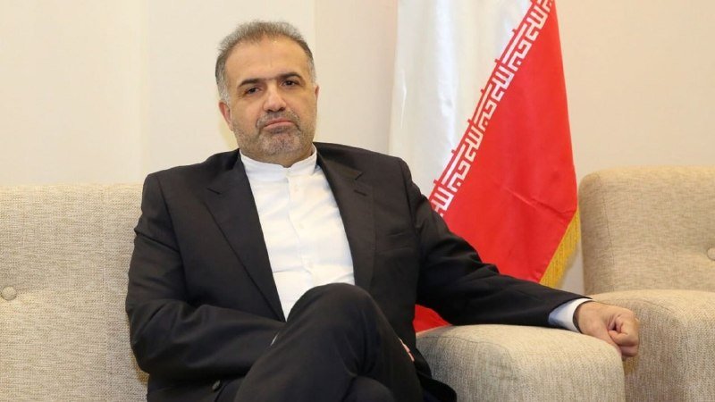 A man in a dark suit and white shirt sits on a beige couch in front of a red flag with a white emblem.